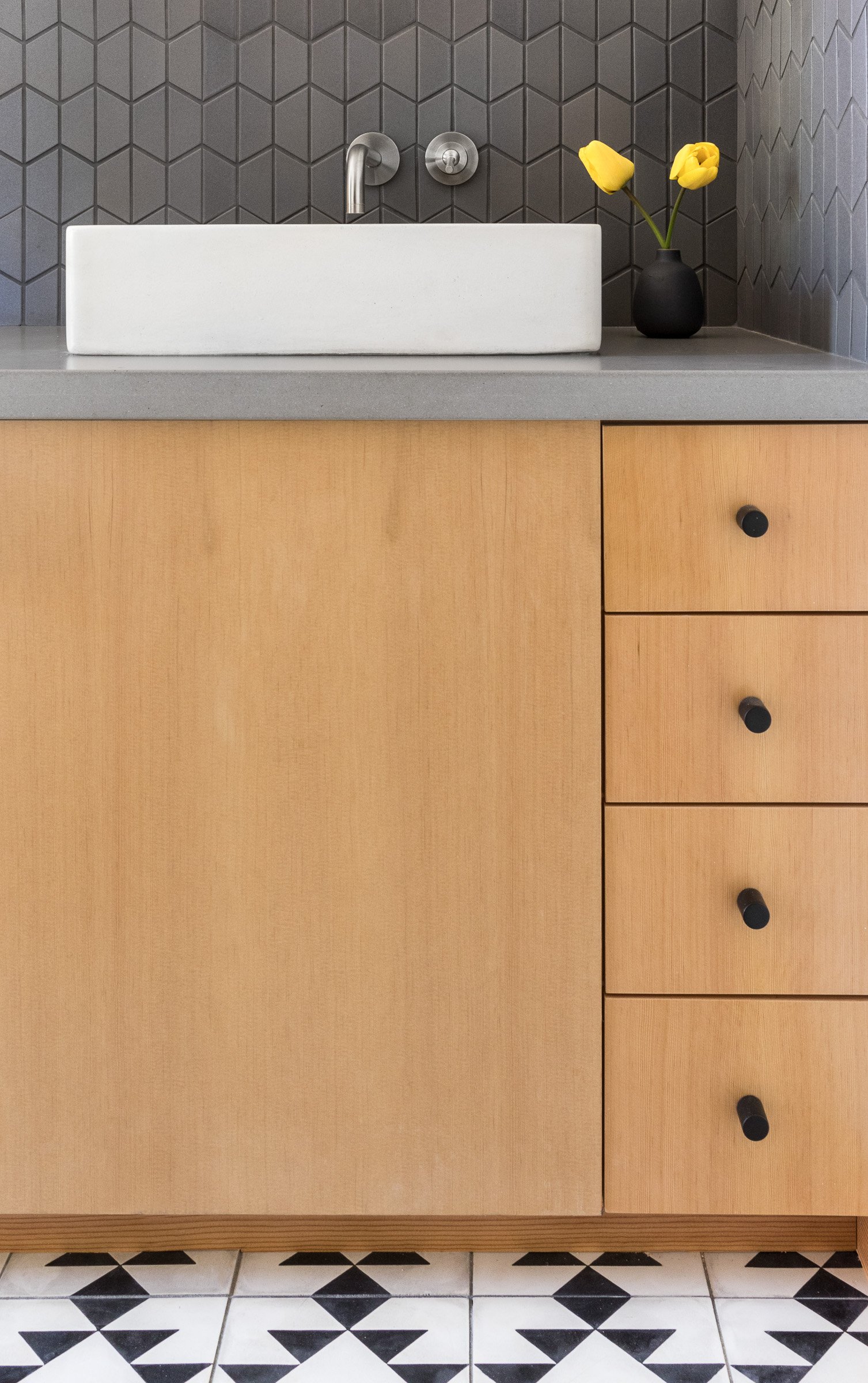 Modern bathroom vanity with wood cabinets, gray countertop, geometric backsplash, white rectangular sink, and black faucet. Yellow flowers in a black vase on the counter.