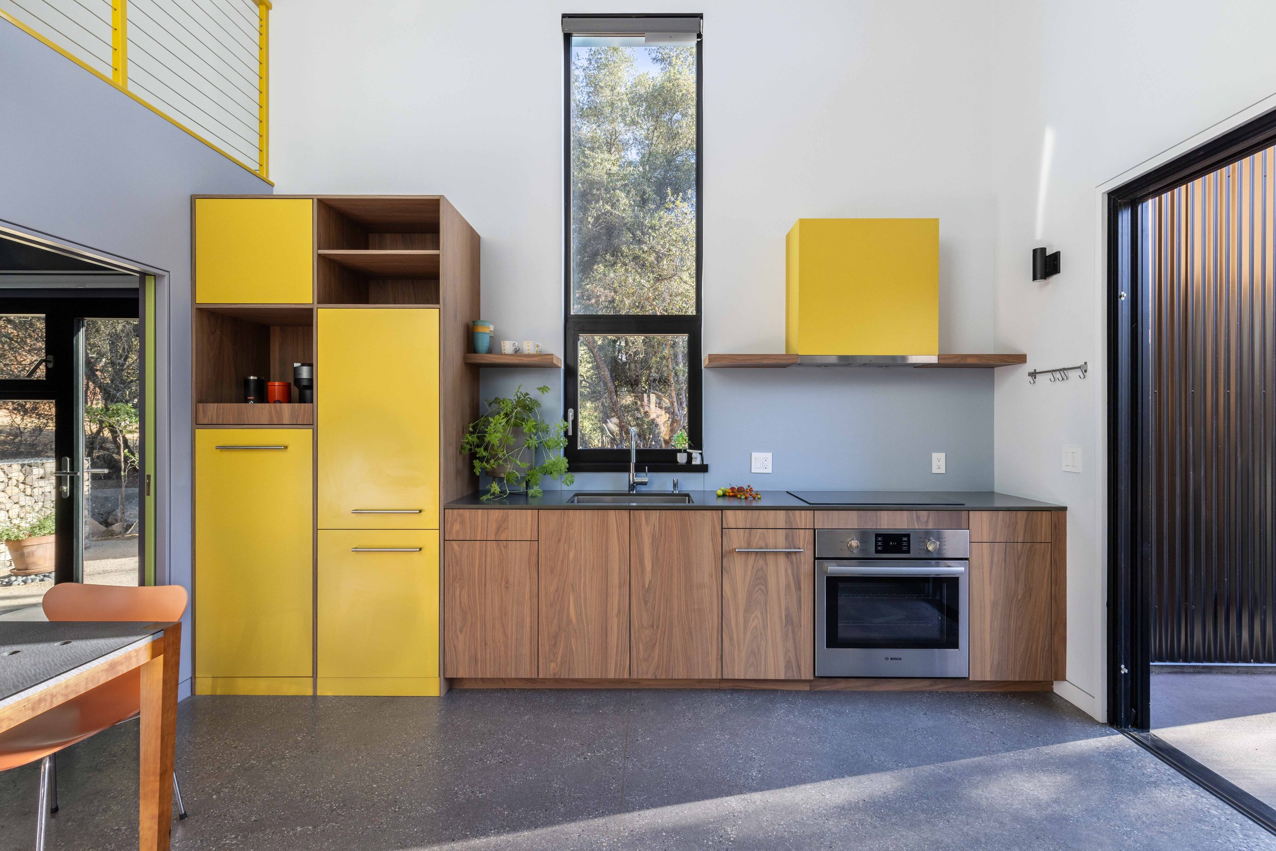 Modern kitchen with yellow cabinets, wood accents, a tall window, built-in oven, and sink under window. Indoor plants and mugs on shelves. Concrete floor.