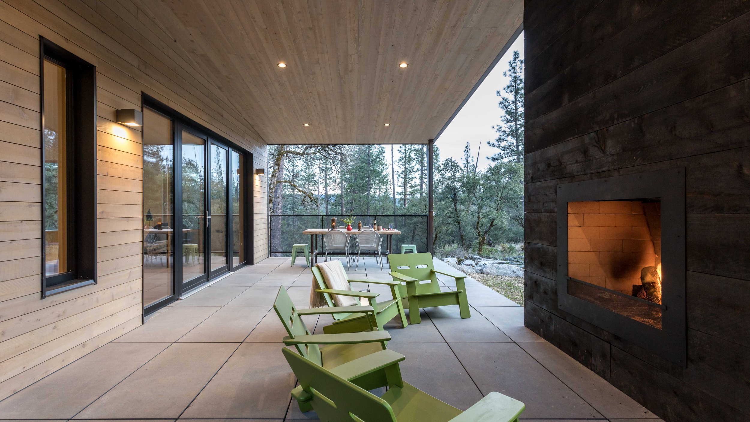Covered patio with green chairs, a dining table, glass doors, and an outdoor fireplace, surrounded by forested landscape.