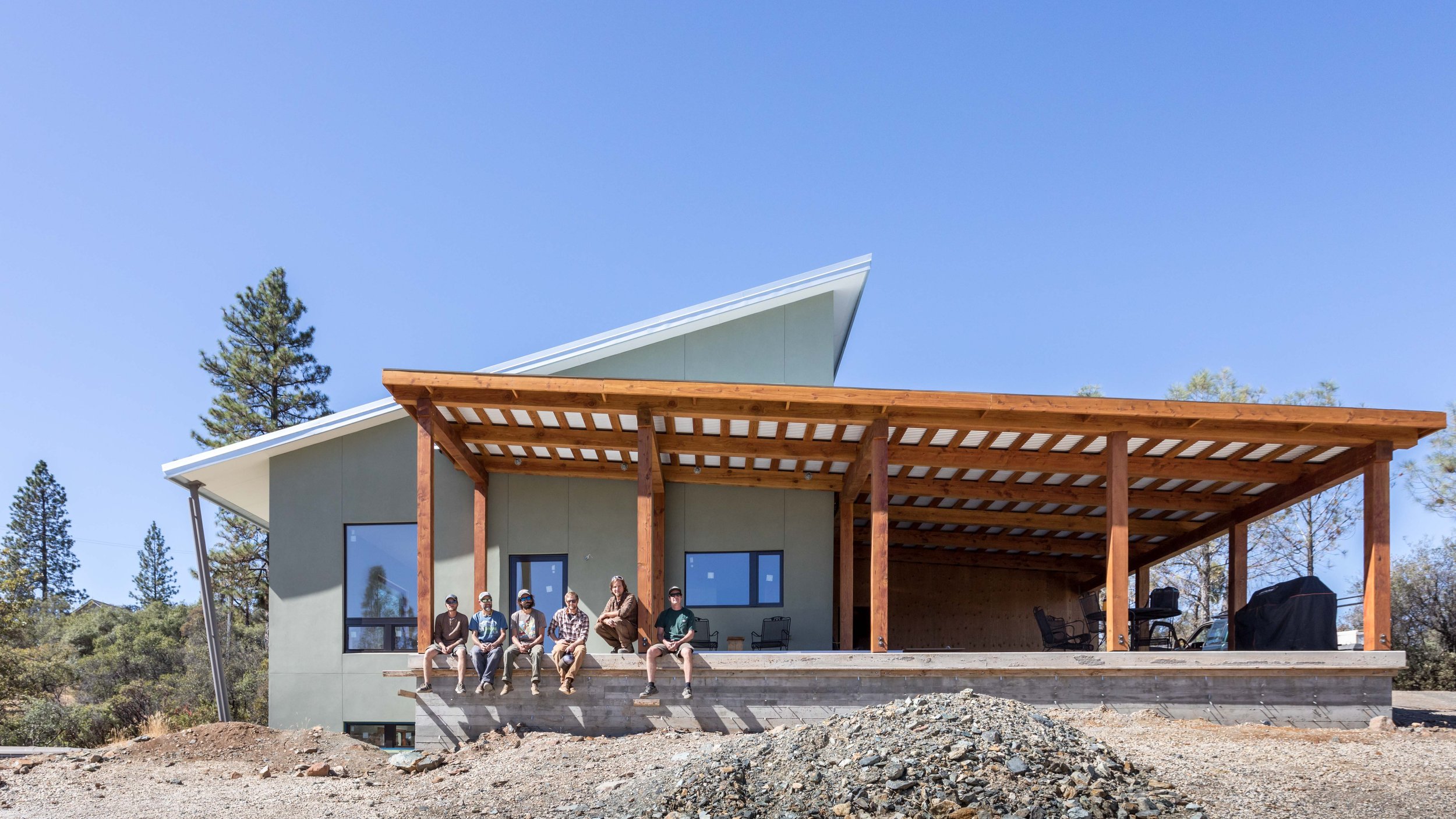 People sit on the porch of a modern house with a slanted roof and wooden pergola. The structure is set in a rural area with trees and a clear sky.