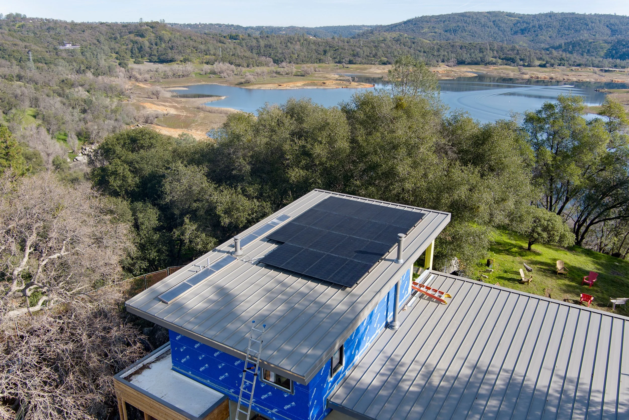 House with solar panels on metal roof, surrounded by trees, overlooking a lake.