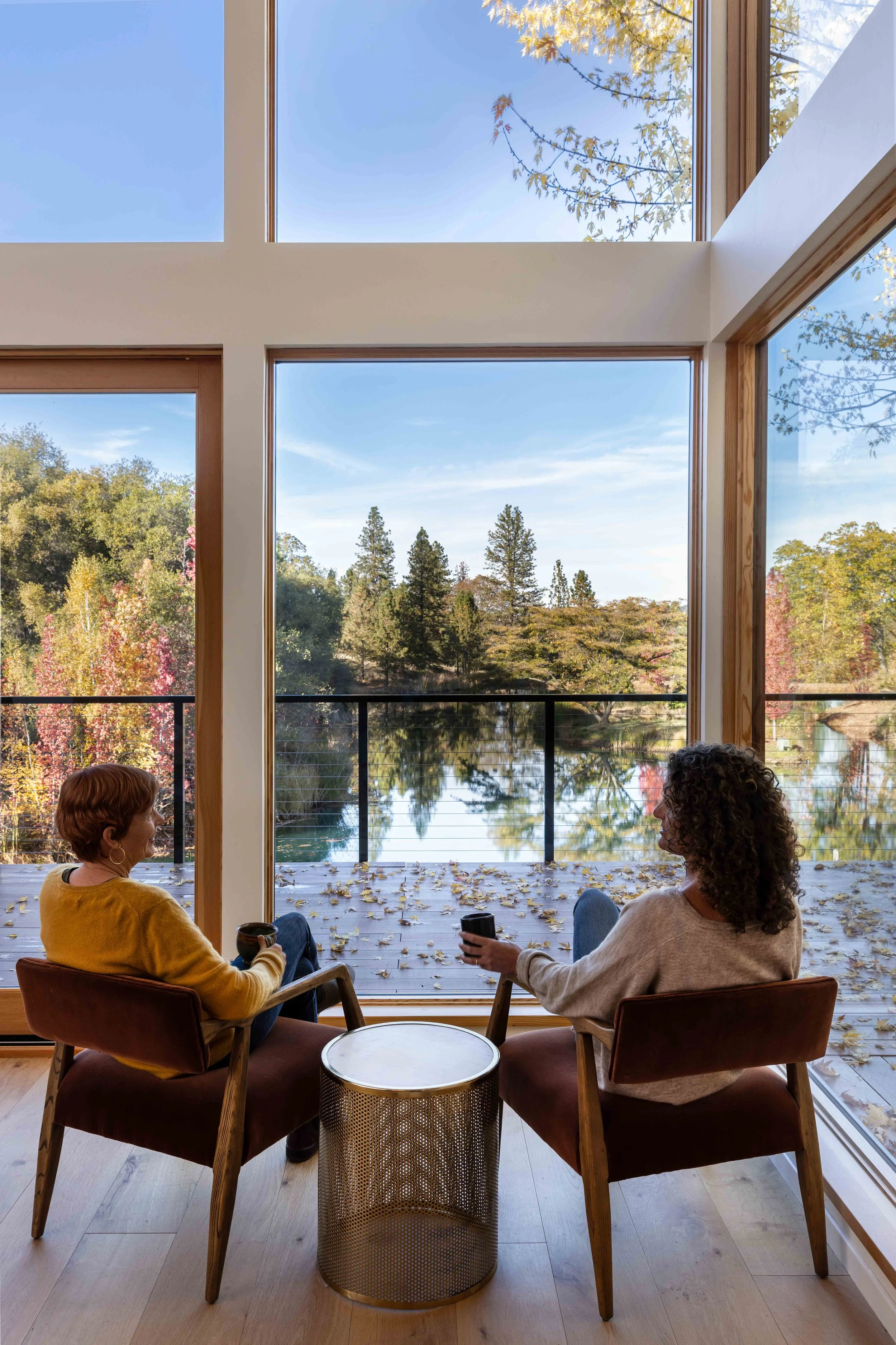 Two people sitting in chairs by large windows, drinking and enjoying a view of trees and a lake.