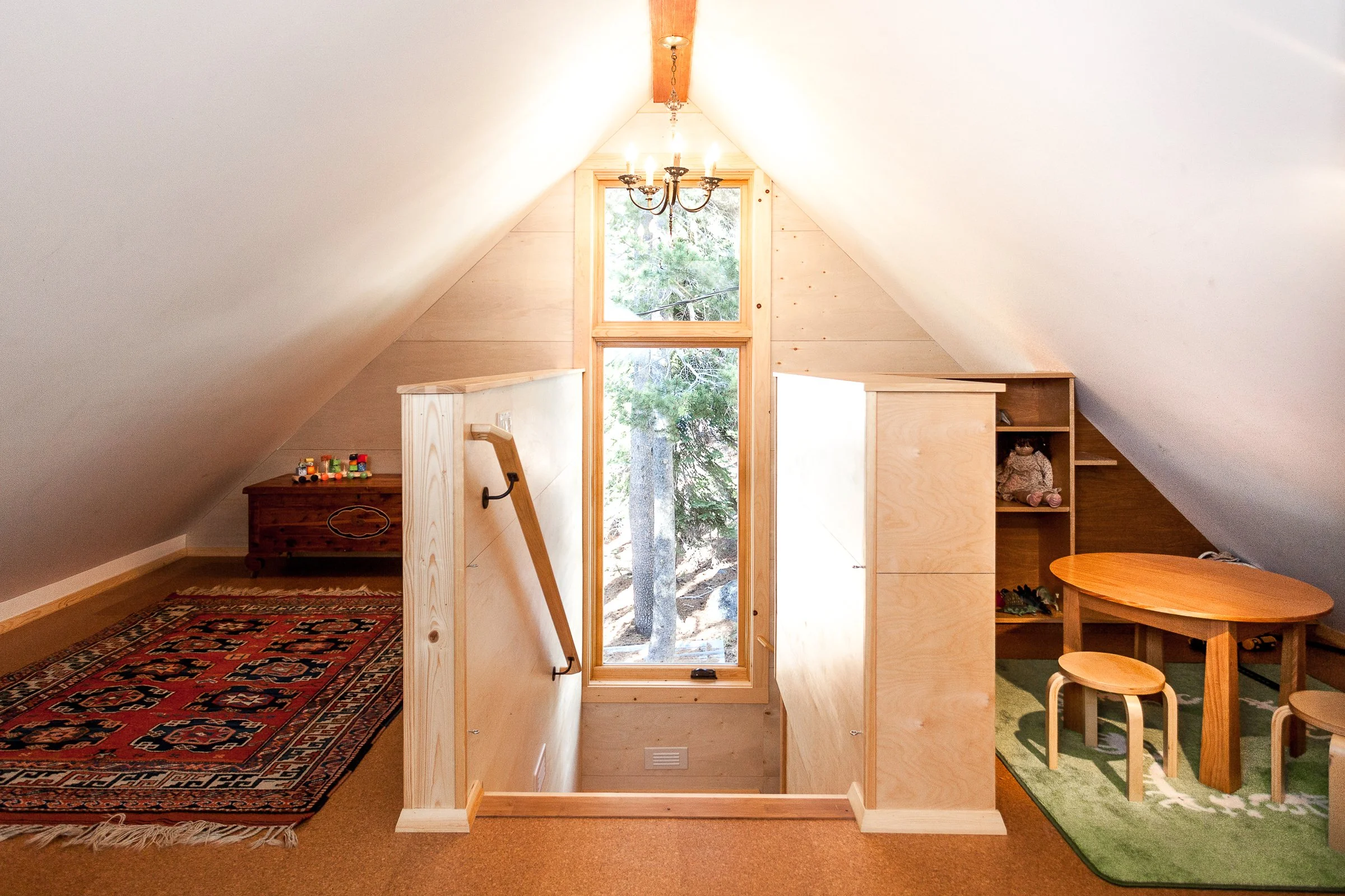 Cozy attic room with a rug, small round table with stools, wooden stairs, large window, chandelier, and wooden shelves.