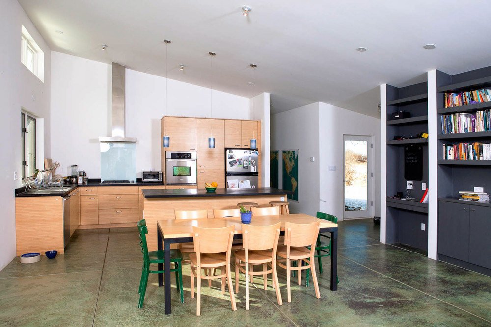 Modern kitchen and dining area with light wood cabinets, a kitchen island, and open shelves. A dining table with chairs is in the center. The floor is concrete, and there are built-in bookshelves on the right.