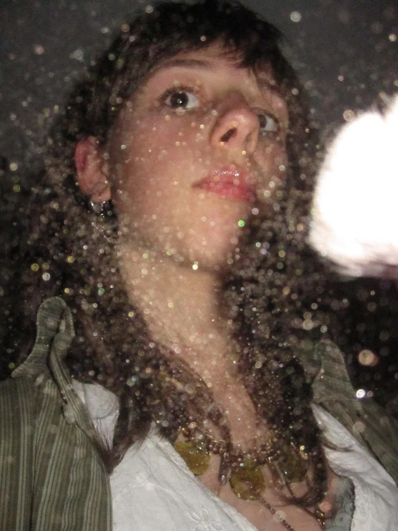  Jasper gazes upwards as sparkles float around her. Light skin, layered brown hair, a white dress, and a green shirt and necklace. Photo courtesy of Clay Lyon. 