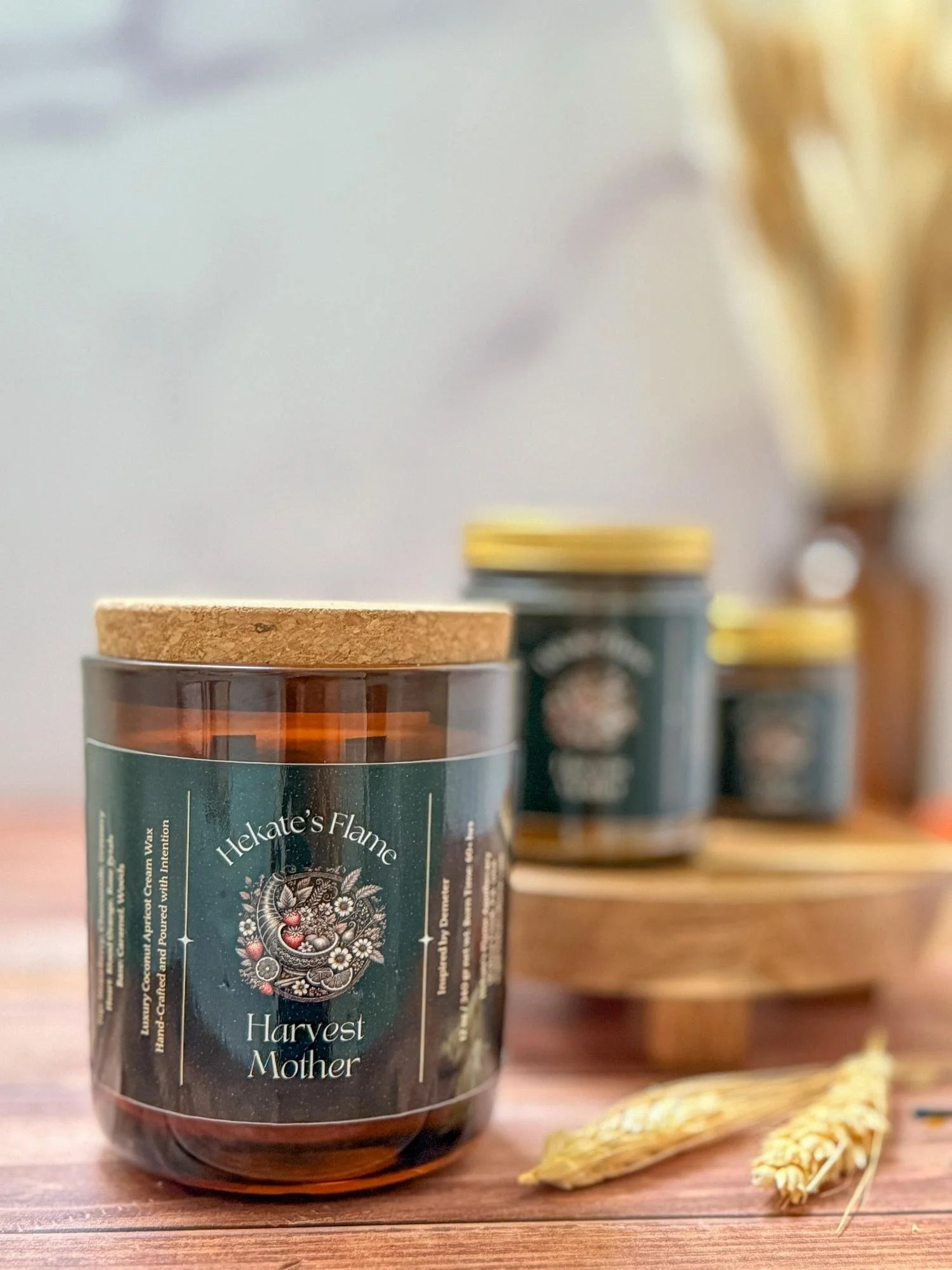 The Harvest Mother candle in a large amber jar with a cork lid is framed by wheat stalks and companion jars in the background.