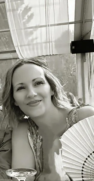 A smiling woman with wavy hair sitting at a table with a fan in front of her inside a room with large windows and a greenhouse ceiling.