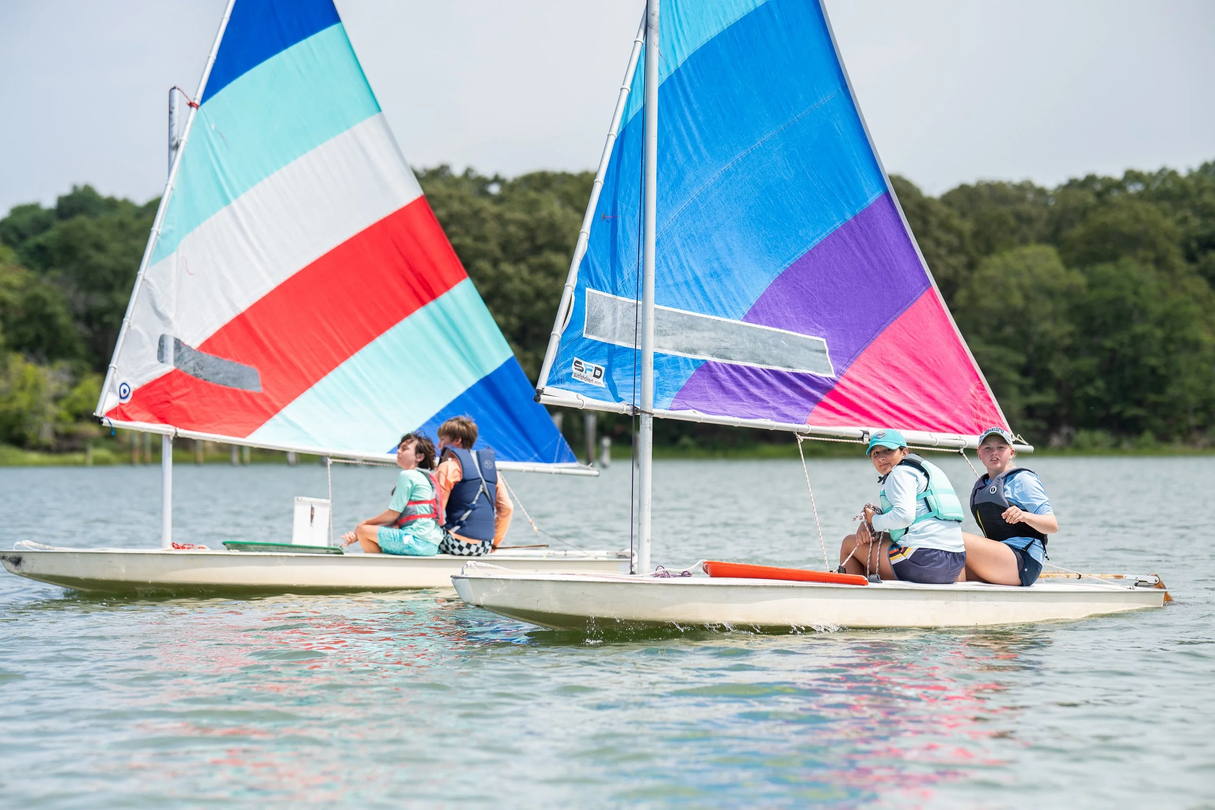 Two young boys sailing a small boat with a colorful sail on a lake, with trees in the background and a sign that reads 'No Wake' on the shore.