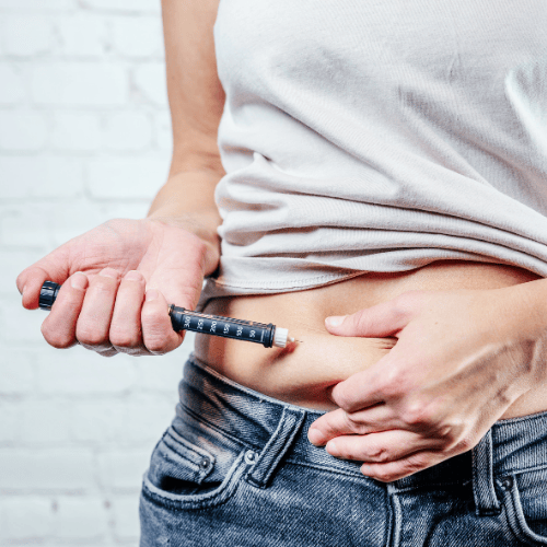A person wearing a white shirt is self-administering an injection into their abdomen with a syringe. The background features a white brick wall.