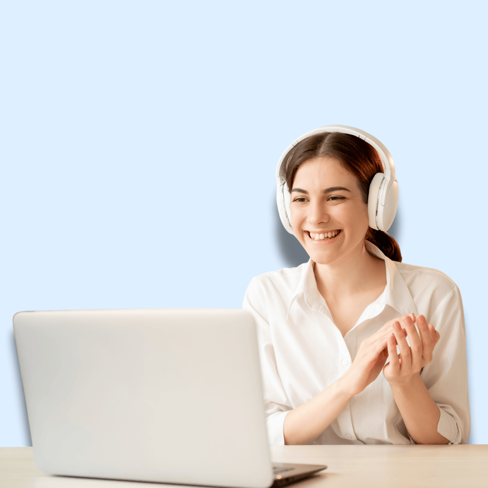 A woman wearing headphones sits at a desk, smiling and clapping her hands while looking at a laptop screen.