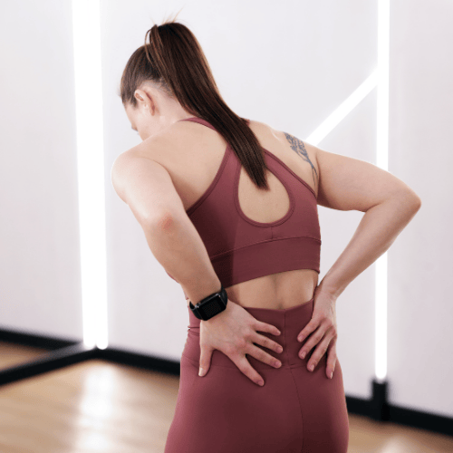 Woman in workout clothes stands indoors, holding her lower back with both hands, possibly experiencing back pain.