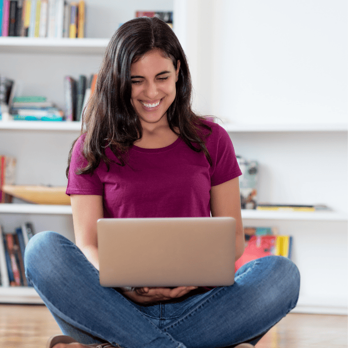 A woman in a purple shirt and jeans sits cross-legged on a floor, smiling as she looks at a laptop. Bookshelves in the background suggest a cozy setting.