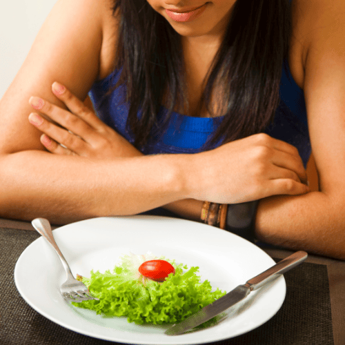 A person sitting at a table looks down at a small salad with a single cherry tomato on a white plate. The mood appears contemplative or dissatisfied.
