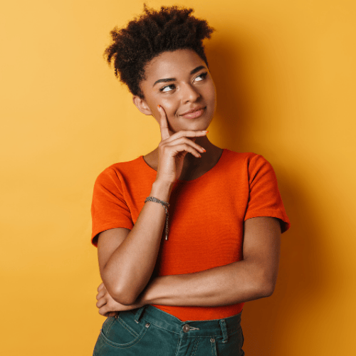 Young person with short curly hair in an orange shirt stands against a yellow background, smiling thoughtfully with a hand on their chin, exuding curiosity.