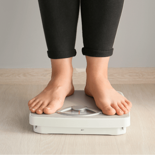 Feet of a person in black pants standing on a white analog bathroom scale, placed on a light wooden floor. The setting conveys a neutral tone.