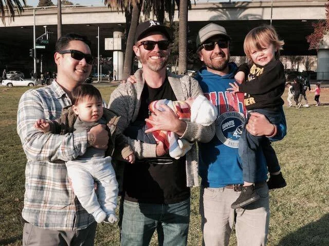 Four adults and two children posing outdoors in a park, with one person holding a wrapped gift.