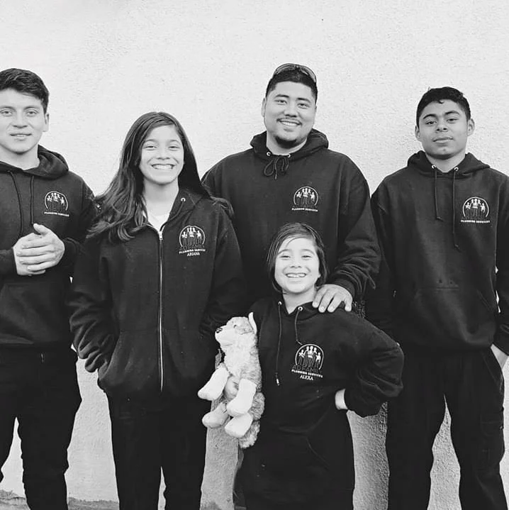 Five young people, four males and one female, standing together in front of a plain wall, all wearing matching black hoodies with a logo, smiling at the camera. The female is holding a stuffed animal.