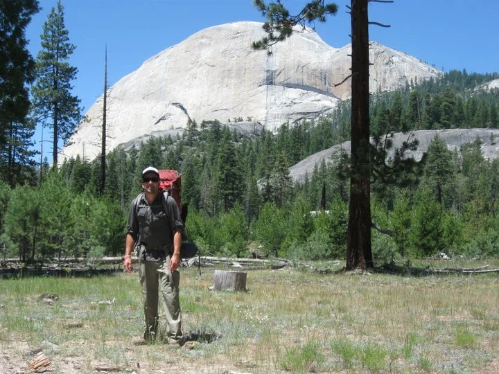 A man with sunglasses, a hat, backpack, and hiking gear standing in a grassy forest clearing with large granite rock formations and pine trees in the background.