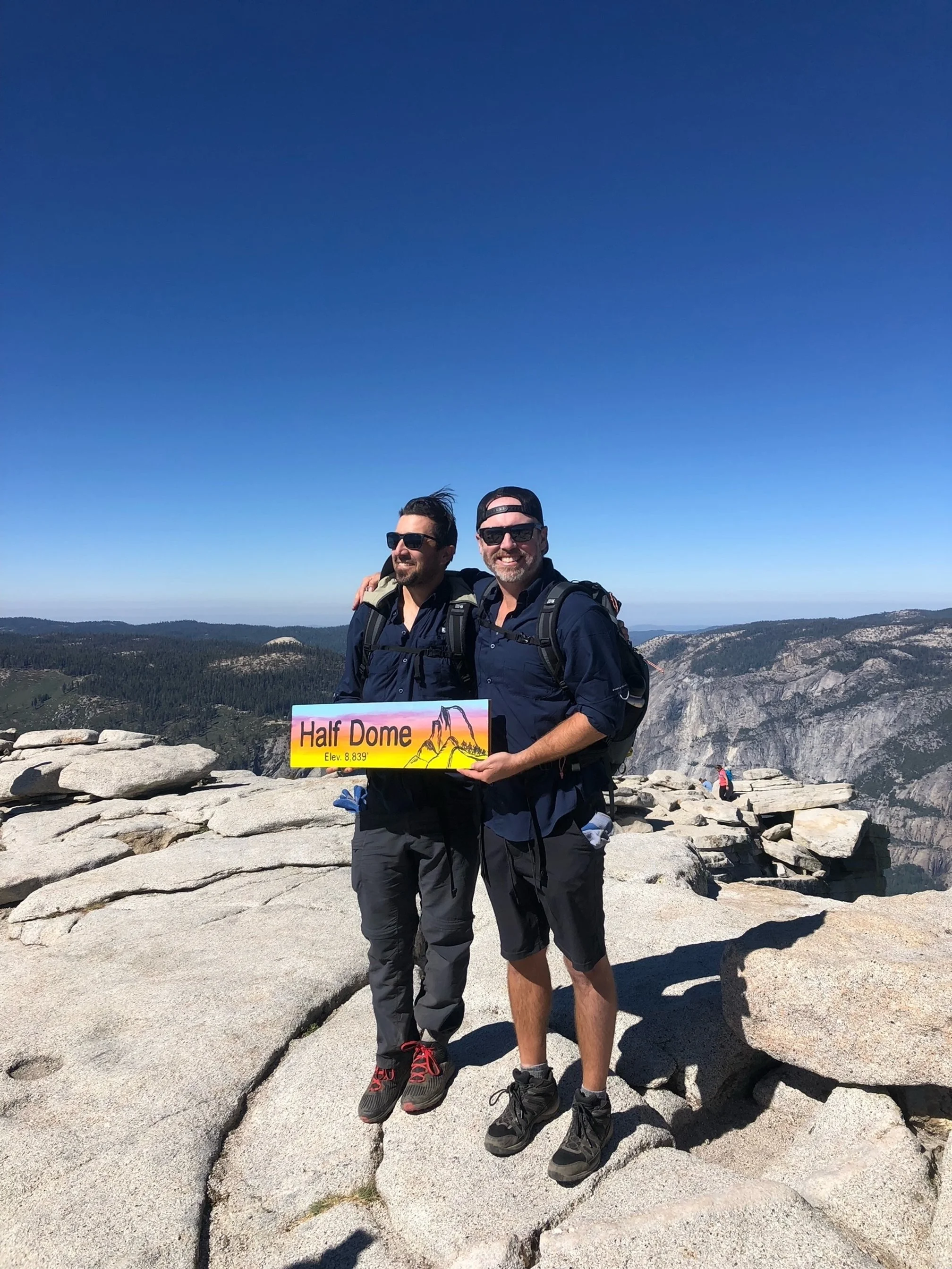 Two men in hiking gear standing on a rocky mountain summit with a sign that reads "Half Dome," smiling with a mountain landscape in the background.