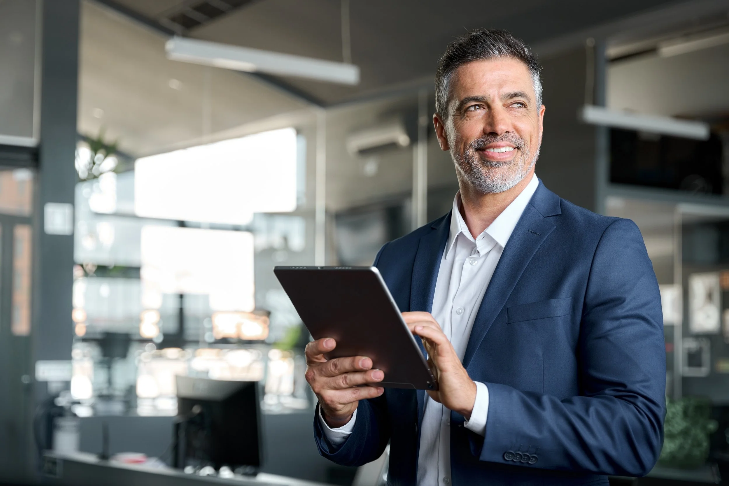 Mature professional man with olive skin and salt-and-pepper hair and beard, dressed in a blue business suit without a tie, holding a tablet and gazing off to the right, standing in a bright, modern office.