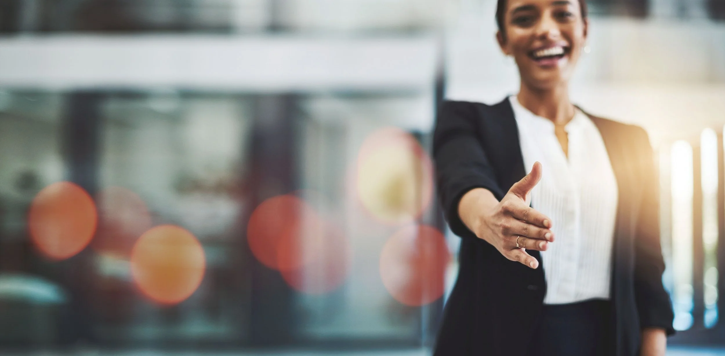 Smiling, dark-haired, woman in a business suit extends right hand for handshake.