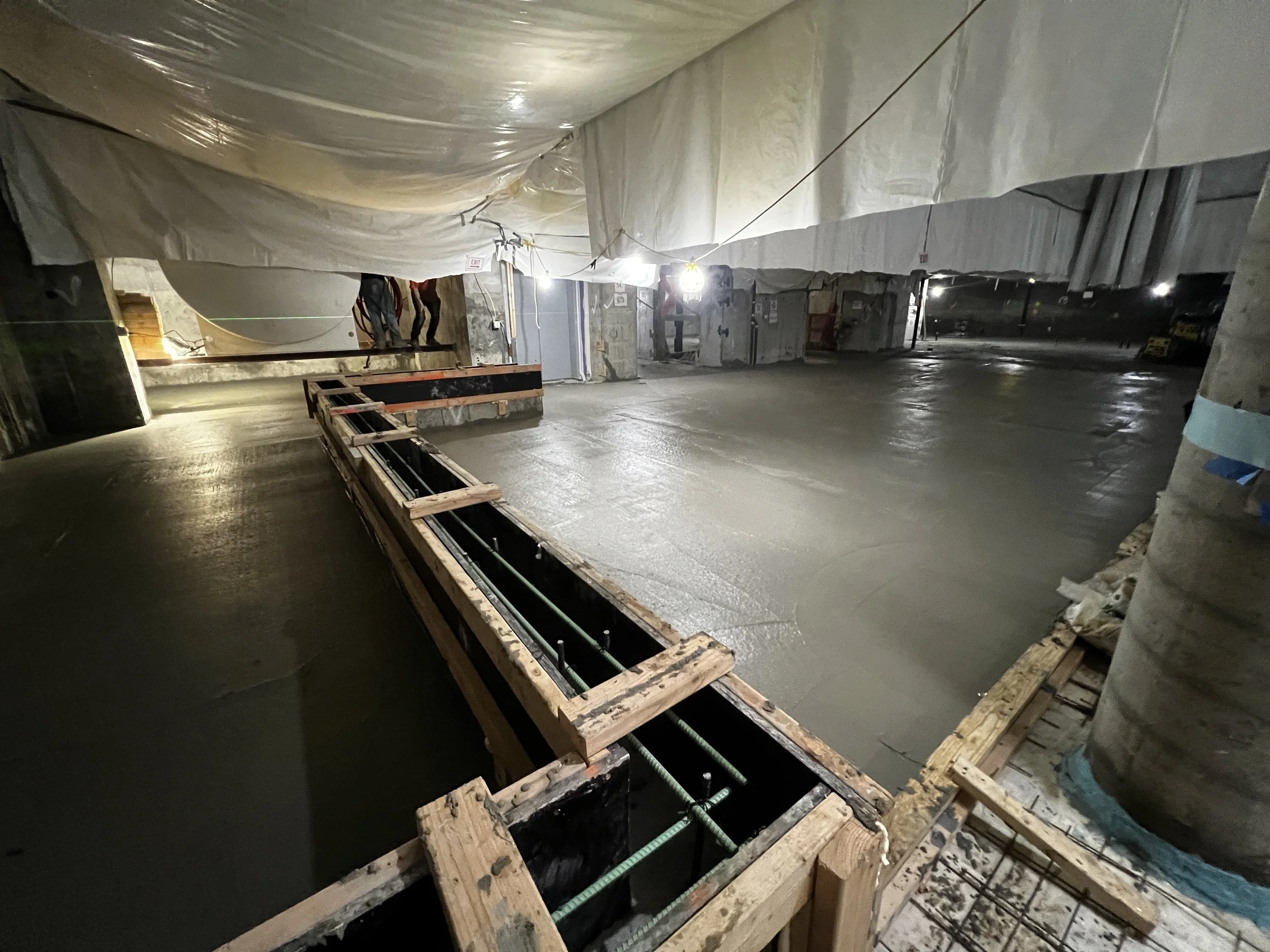 Construction site with a large area of freshly poured concrete. Wooden forms are in place, and safety barriers are visible. Workers can be seen in the background, and the area is illuminated by construction lights.