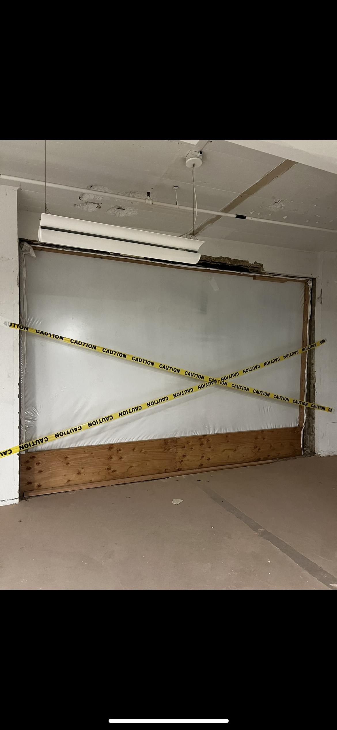 A garage door is covered with a white protective sheet and caution tape, with some exposed wood at the bottom. The area appears to be under construction or renovation.