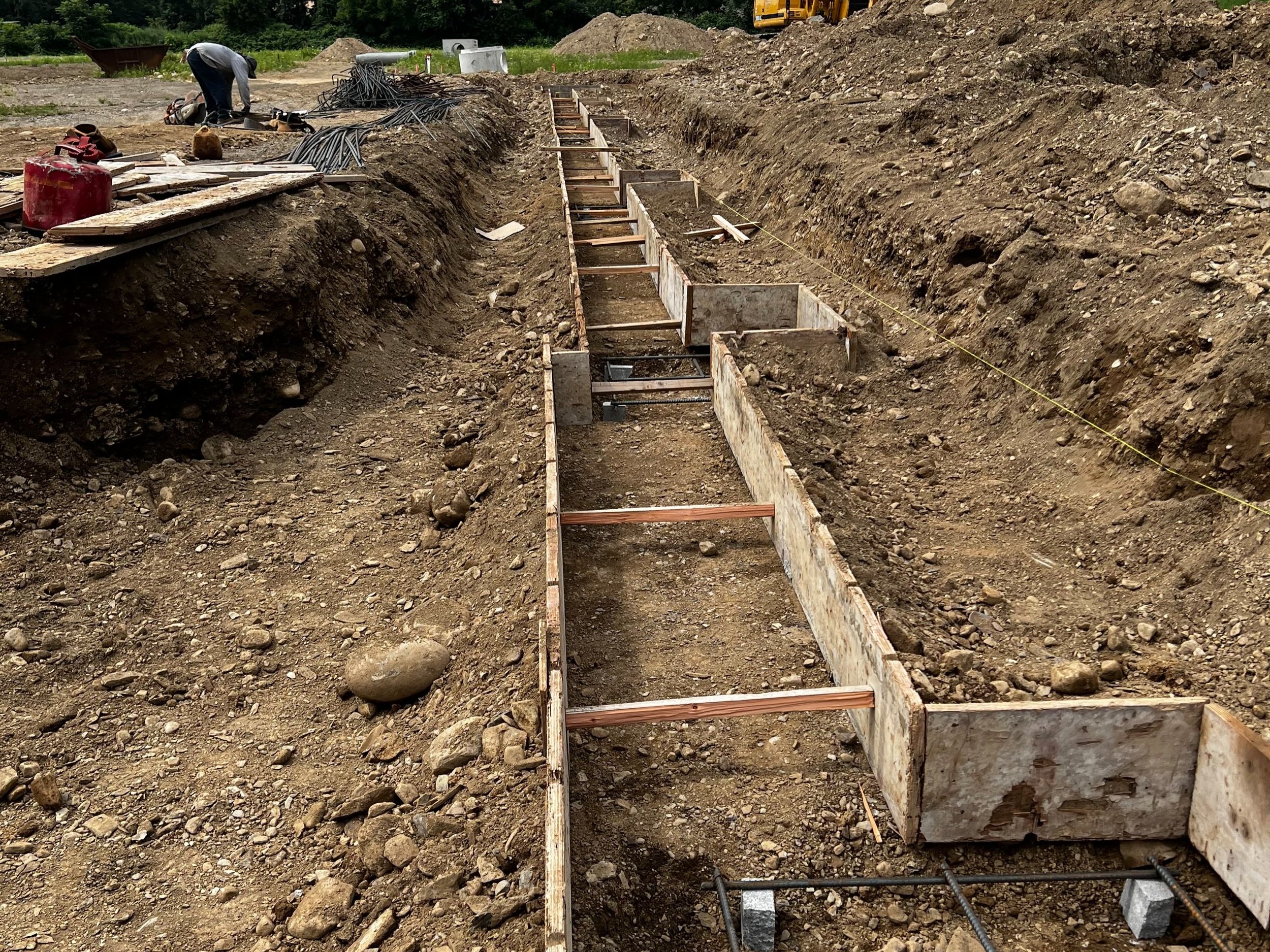Construction site with a long rectangular trench reinforced with wooden and metal supports, and a worker in the background working on the site.
