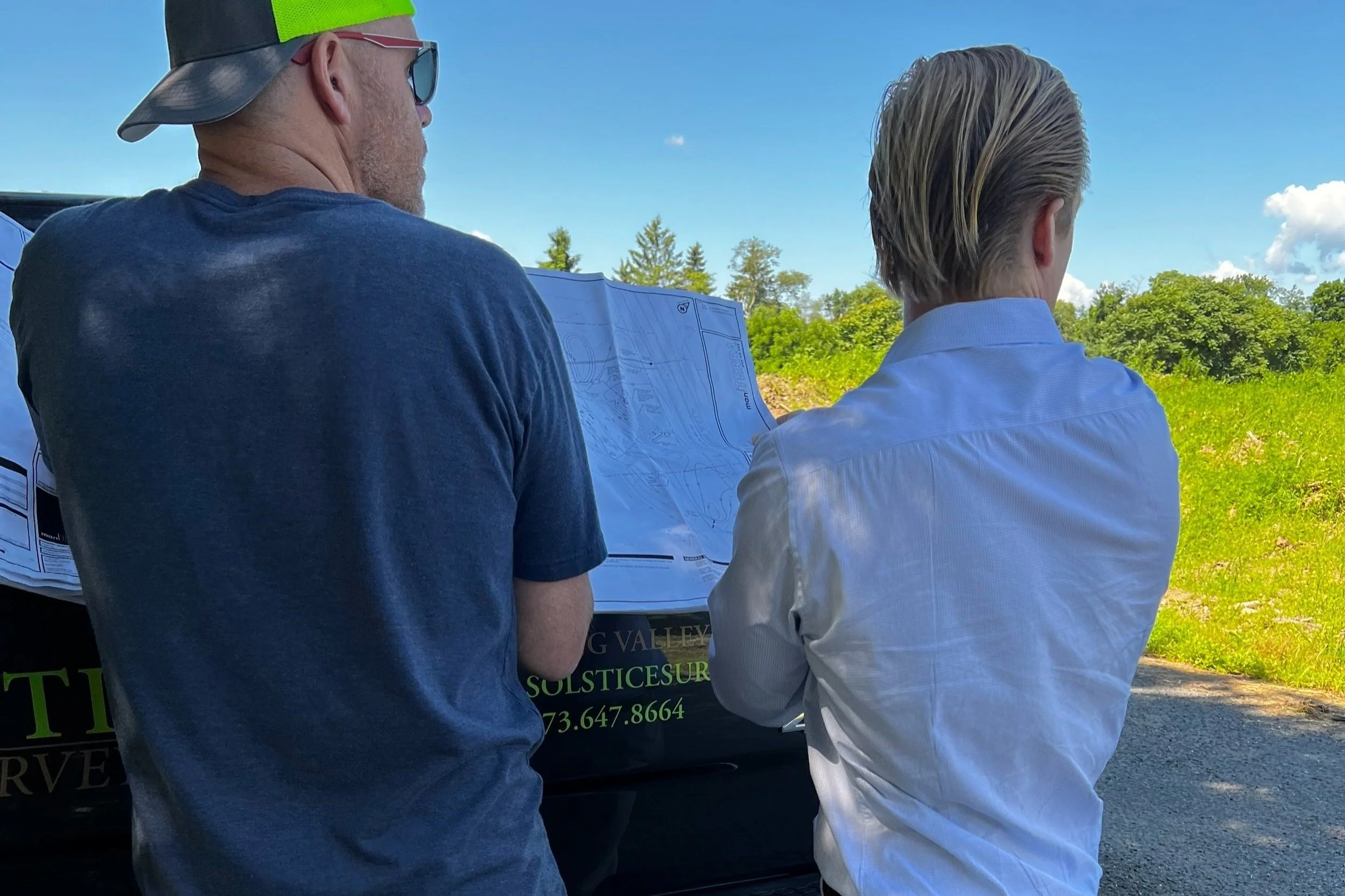 Two men look at plans or blueprints outdoors on a sunny day, with green trees and grass in the background.