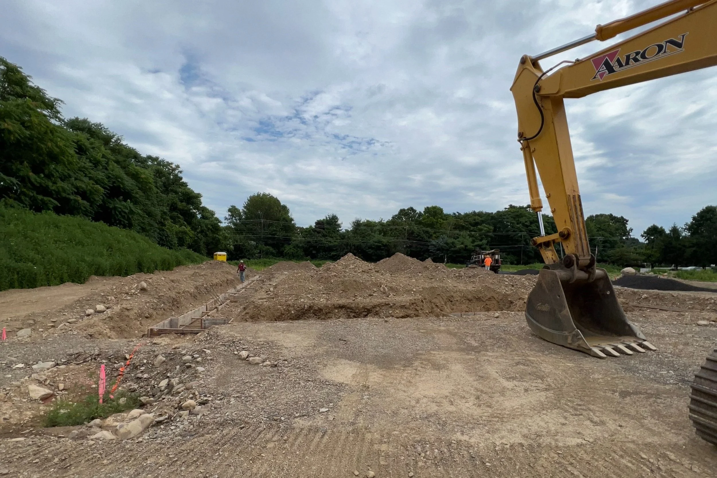 Construction site with excavator and workers, dirt piles, and open blue sky.