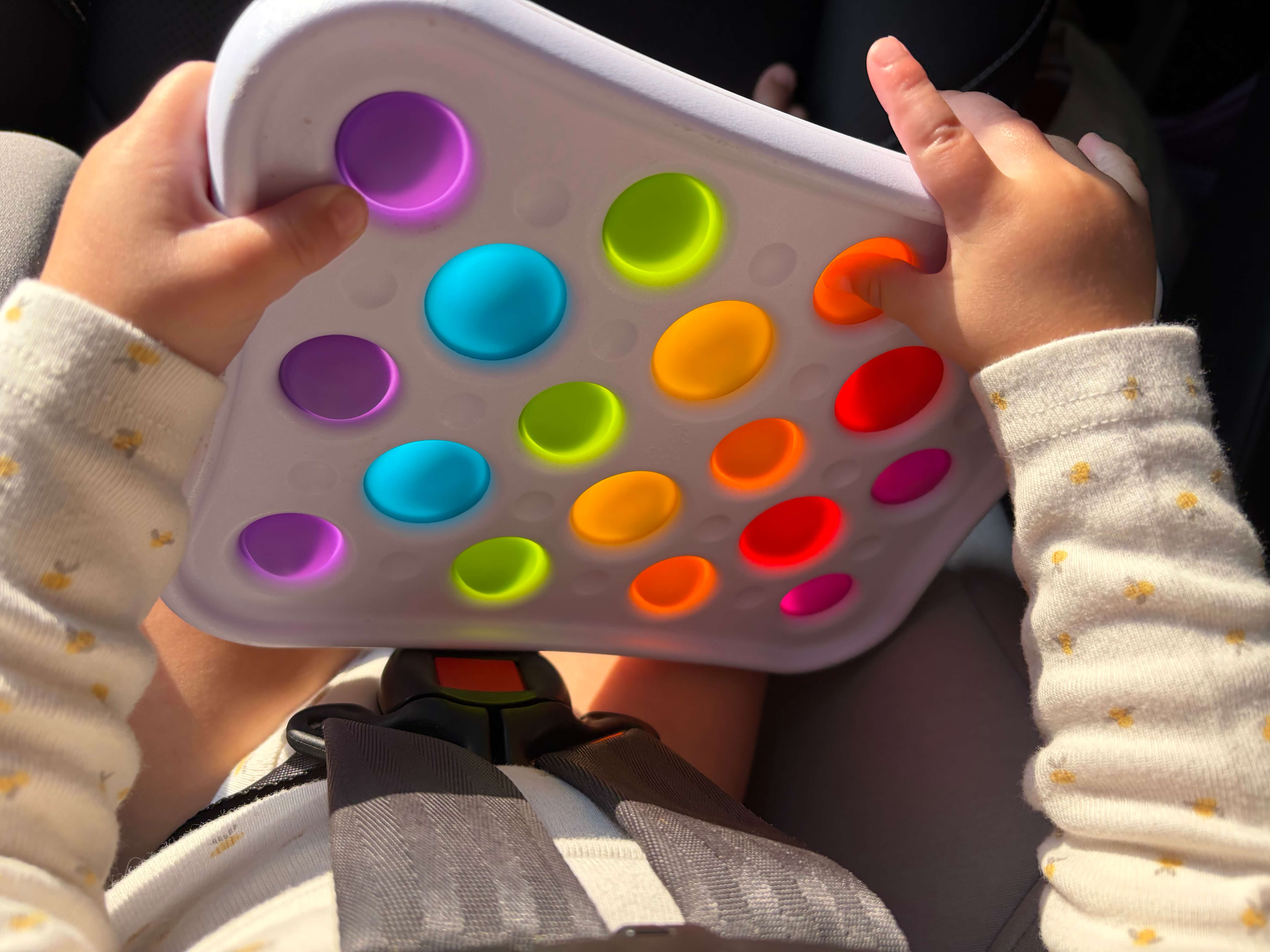toddler holding a fat brain toys dimpl popper toy while traveling in a carseat