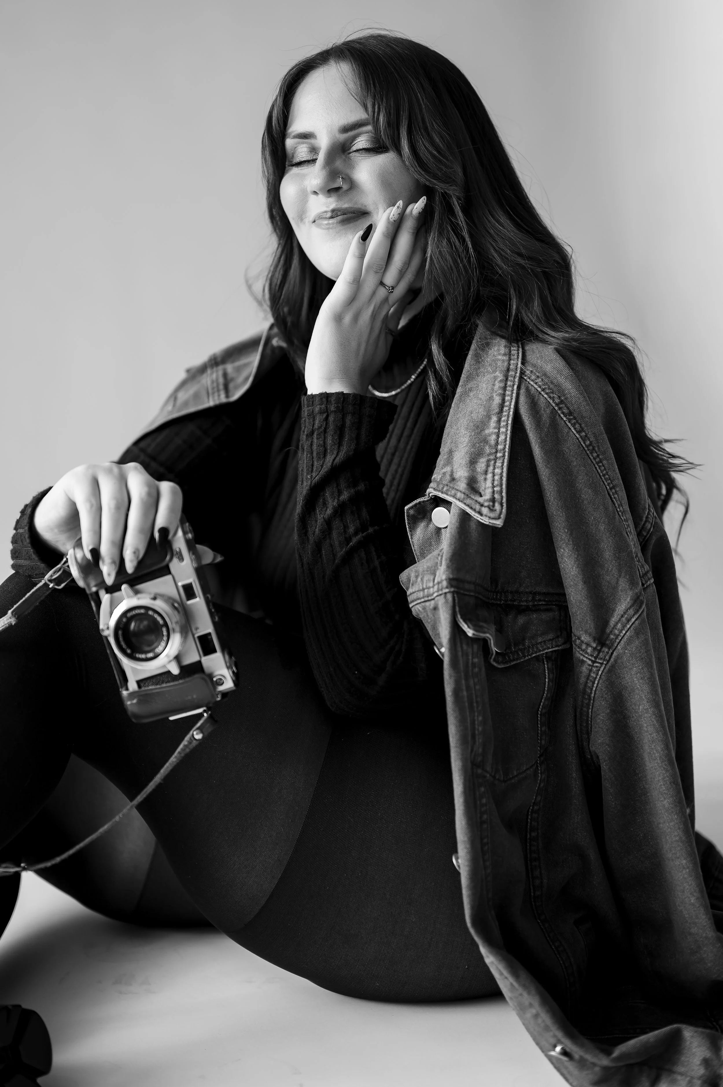 A woman sitting on the floor with a vintage camera in her hand, smiling with her eyes closed, wearing a dark outfit and a denim jacket draped over her shoulder.