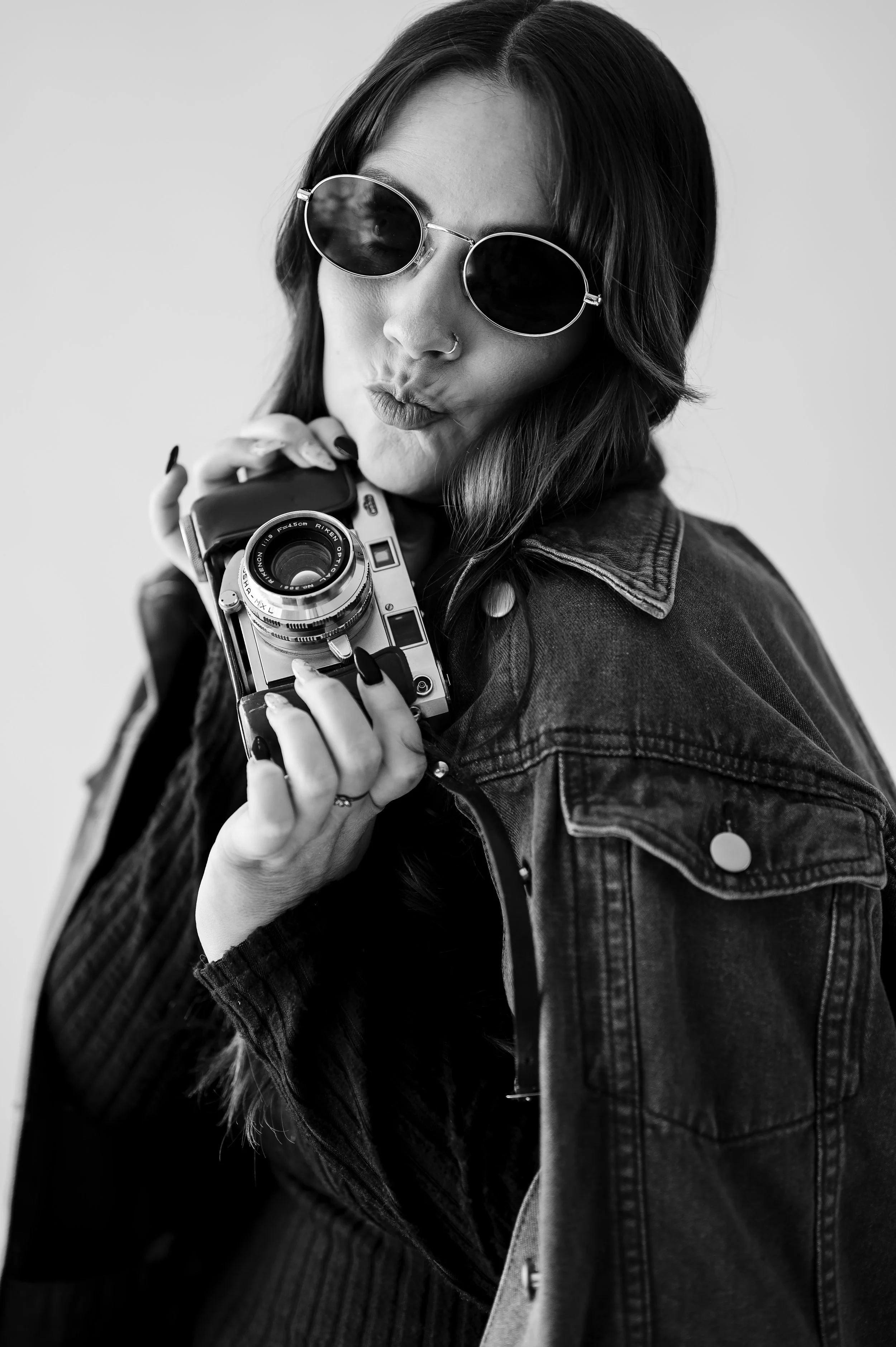 Black-and-white photo of a woman with long dark hair wearing sunglasses and a denim jacket, holding a vintage camera near her face, making a playful puckered-lips expression.