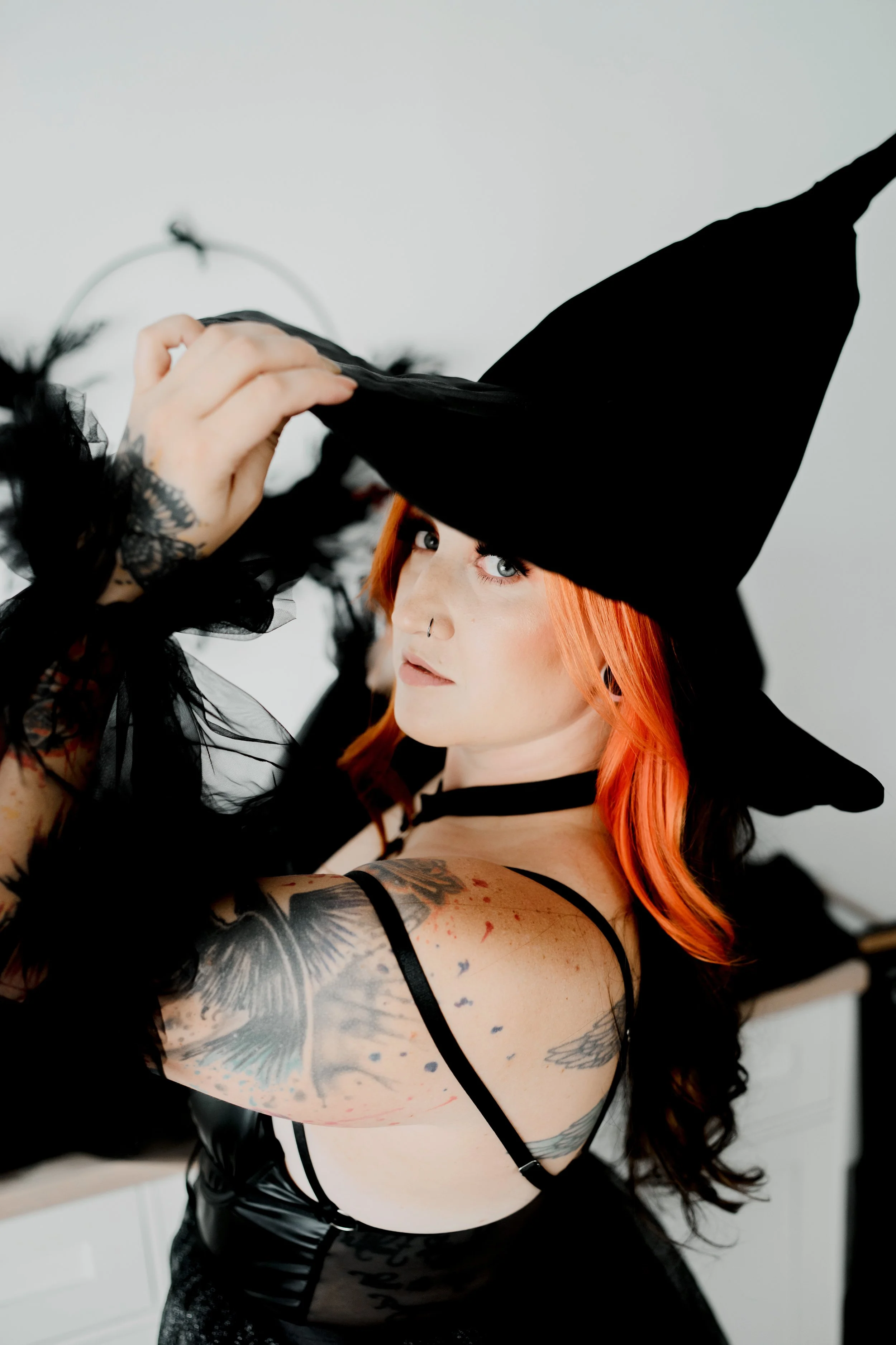Woman dressed as a witch in a black hat, with tattoos and orange hair, adjusting her hat