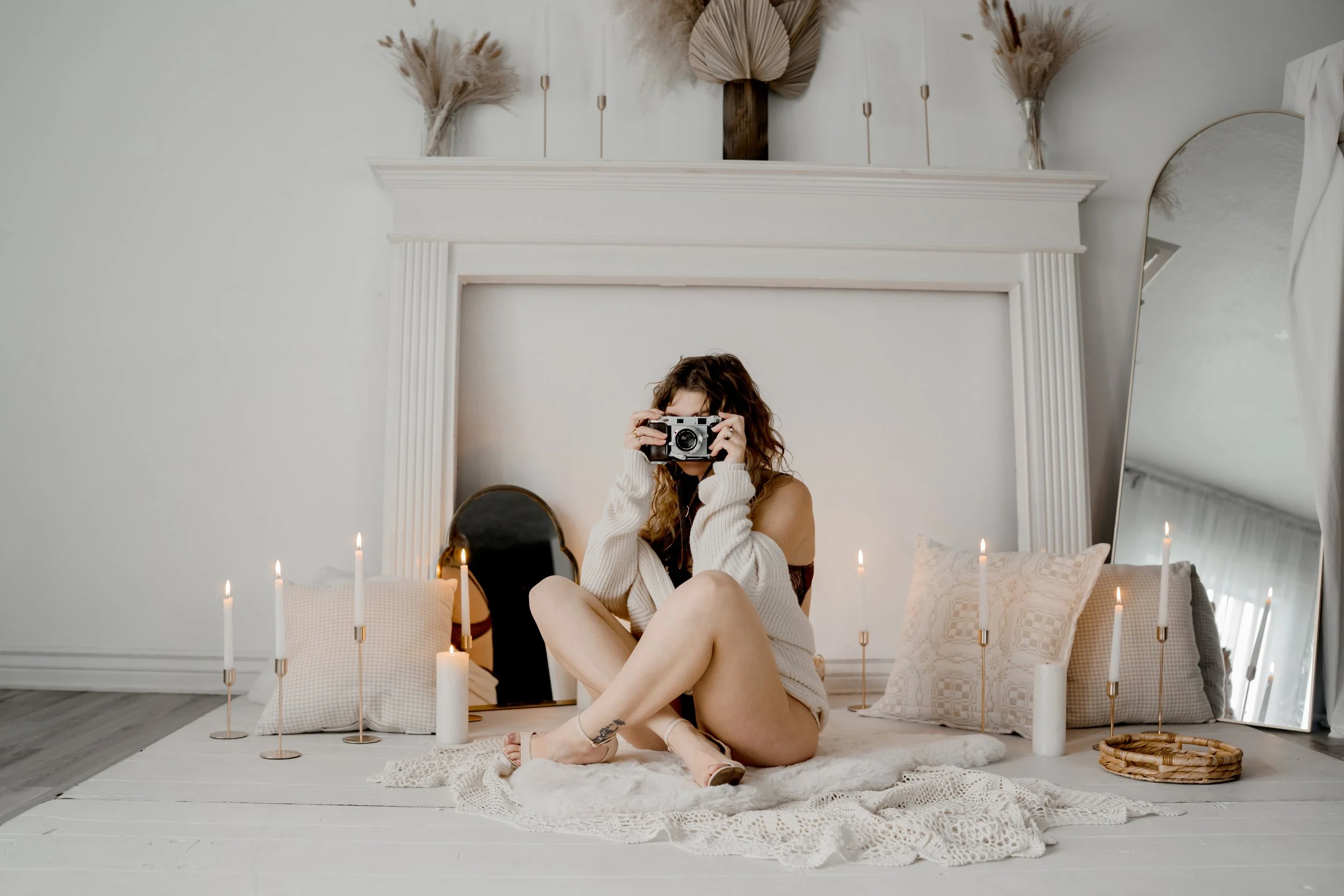 A woman sitting on a white rug in front of a white fireplace, holding a vintage camera to her face and taking a photo. The room is decorated with candles, pillows, a standing mirror, and dried floral arrangements on the mantel.