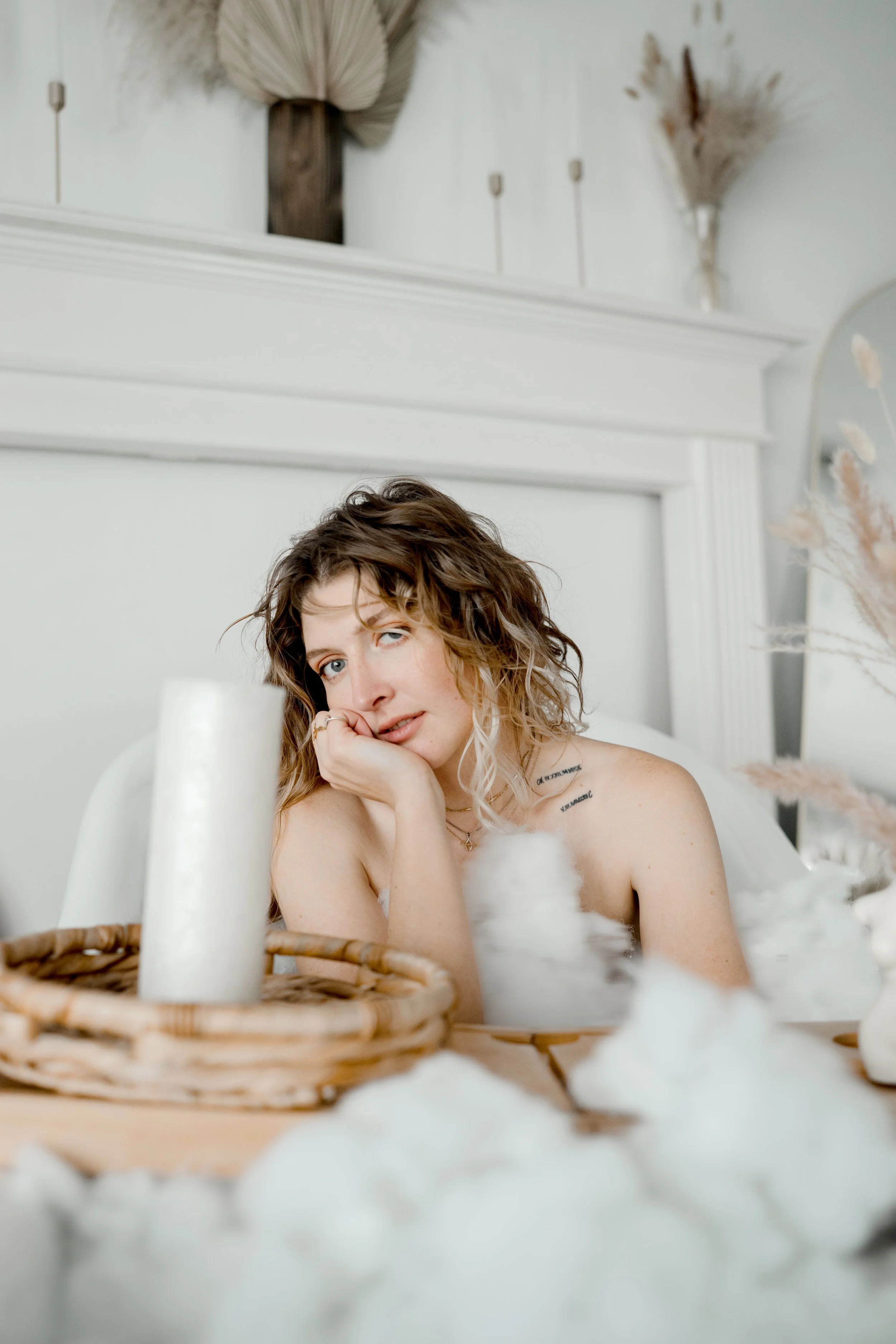 A woman with wavy brown hair and blue eyes relaxing in a bubble bath, resting her chin on her hand with a gentle expression. The bathroom has a white aesthetic with dried floral decorations and a basket with a white candle on a wooden tray in the for