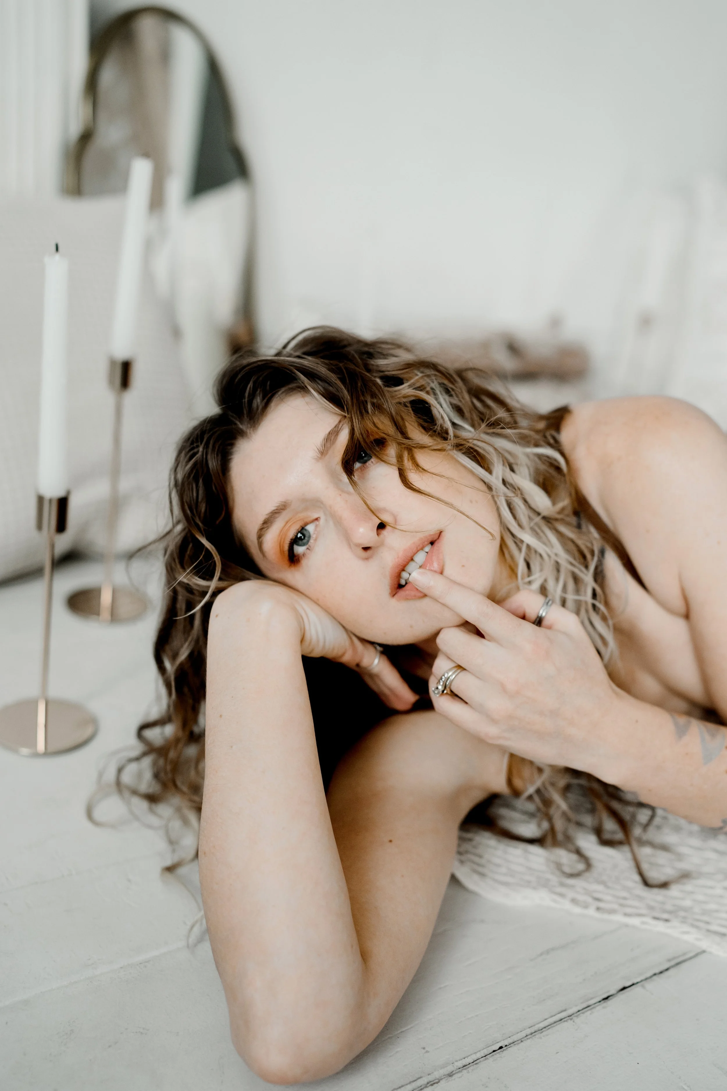 A woman with curly hair lying on the floor, propped on her arm, with her finger near her lips, looking contemplative or sensual.