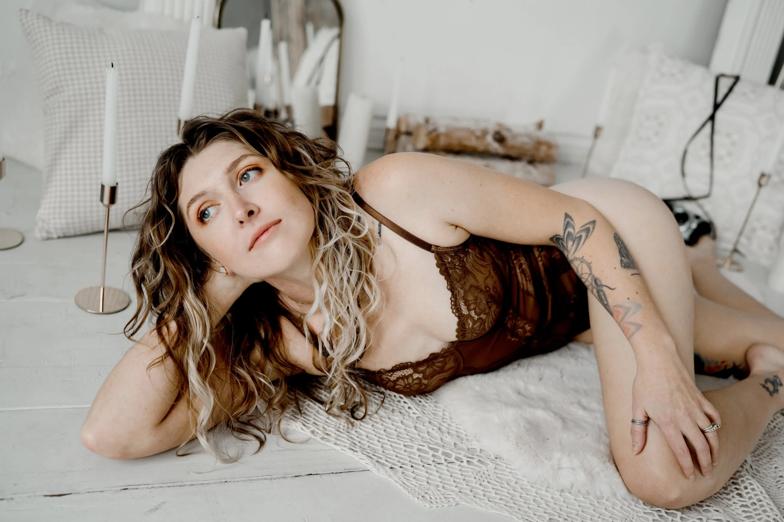 A woman with wavy blonde hair and tattoos lying on a bed, looking thoughtfully to the side, in a room with candles, pillows, and wooden furniture.