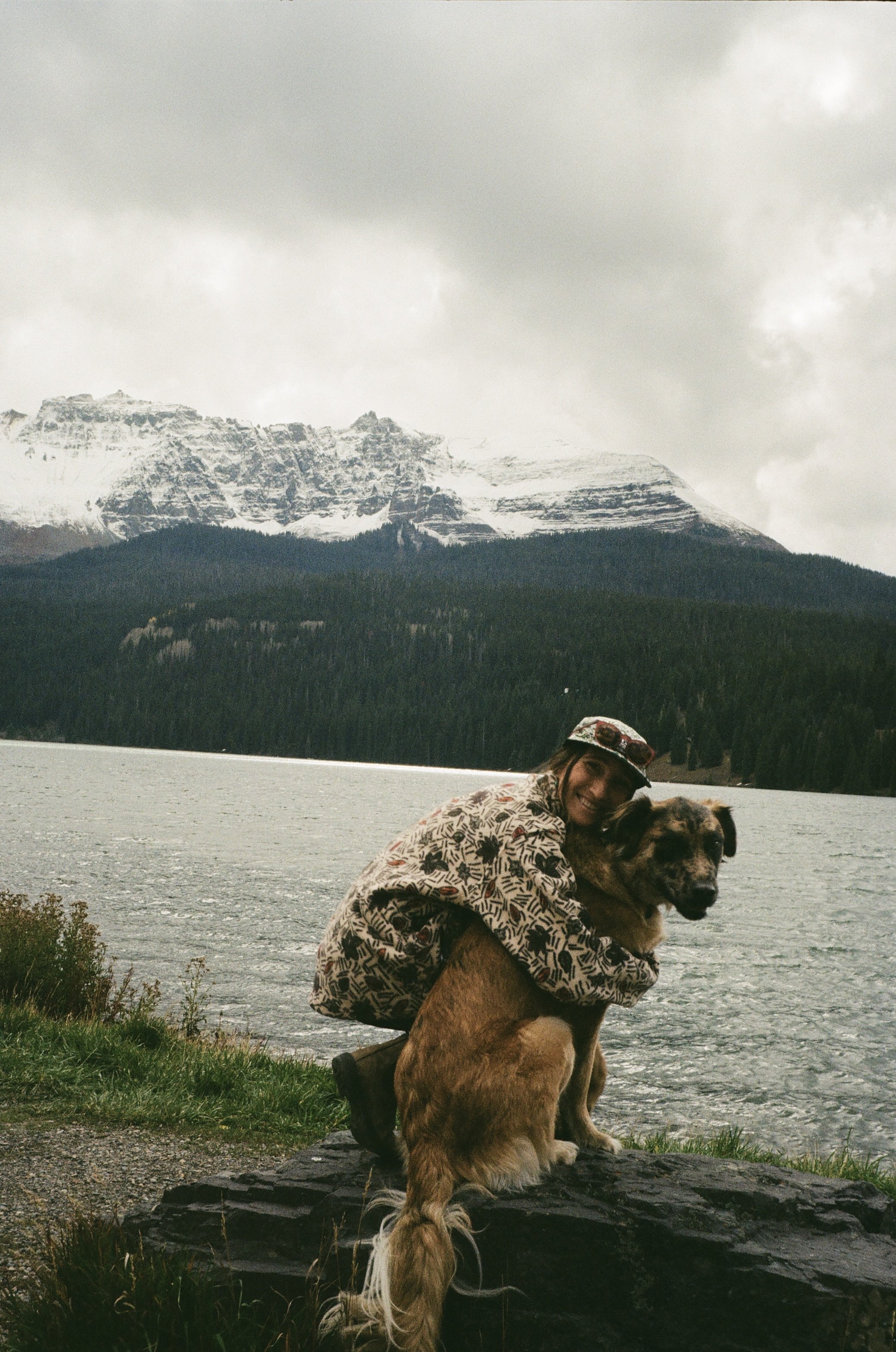 A woman in a patterned jacket and a cap hugging a large dog with a snow-capped mountain and cloudy sky in the background, next to a body of water.