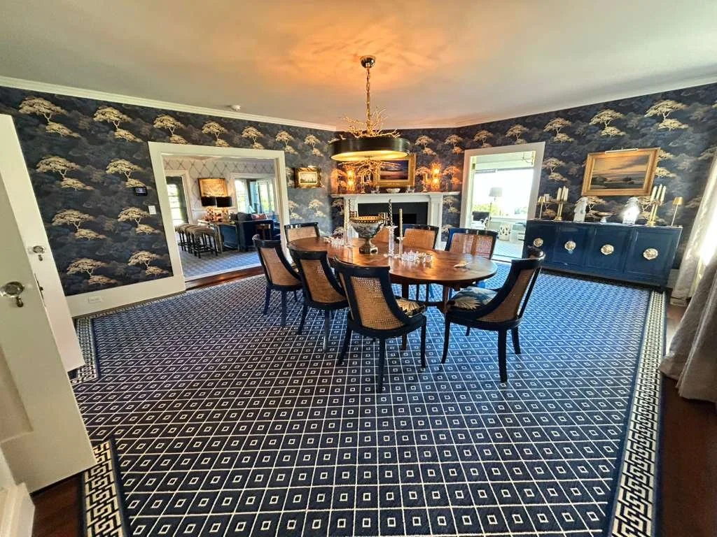 A dining room with a large wooden table and eight chairs. The room has blue wallpaper with a cloud-like pattern, a dark blue sideboard with gold accents, and artwork on the walls. There is a window on one side and a doorway leading to a living room w