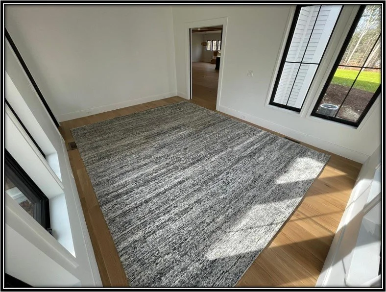 Empty room with large gray rug, light wood floor, white walls, and large windows letting in natural light.