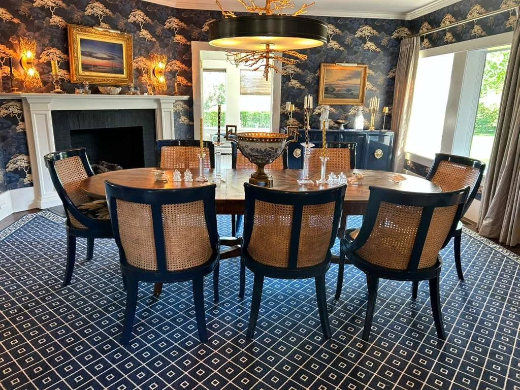 A dining room with a wooden table and eight chairs with woven cane backs, set with glassware and candles. The room has a blue and gold wallpaper, a fireplace with a white mantel, and a large window with curtains, allowing natural light to illuminate 