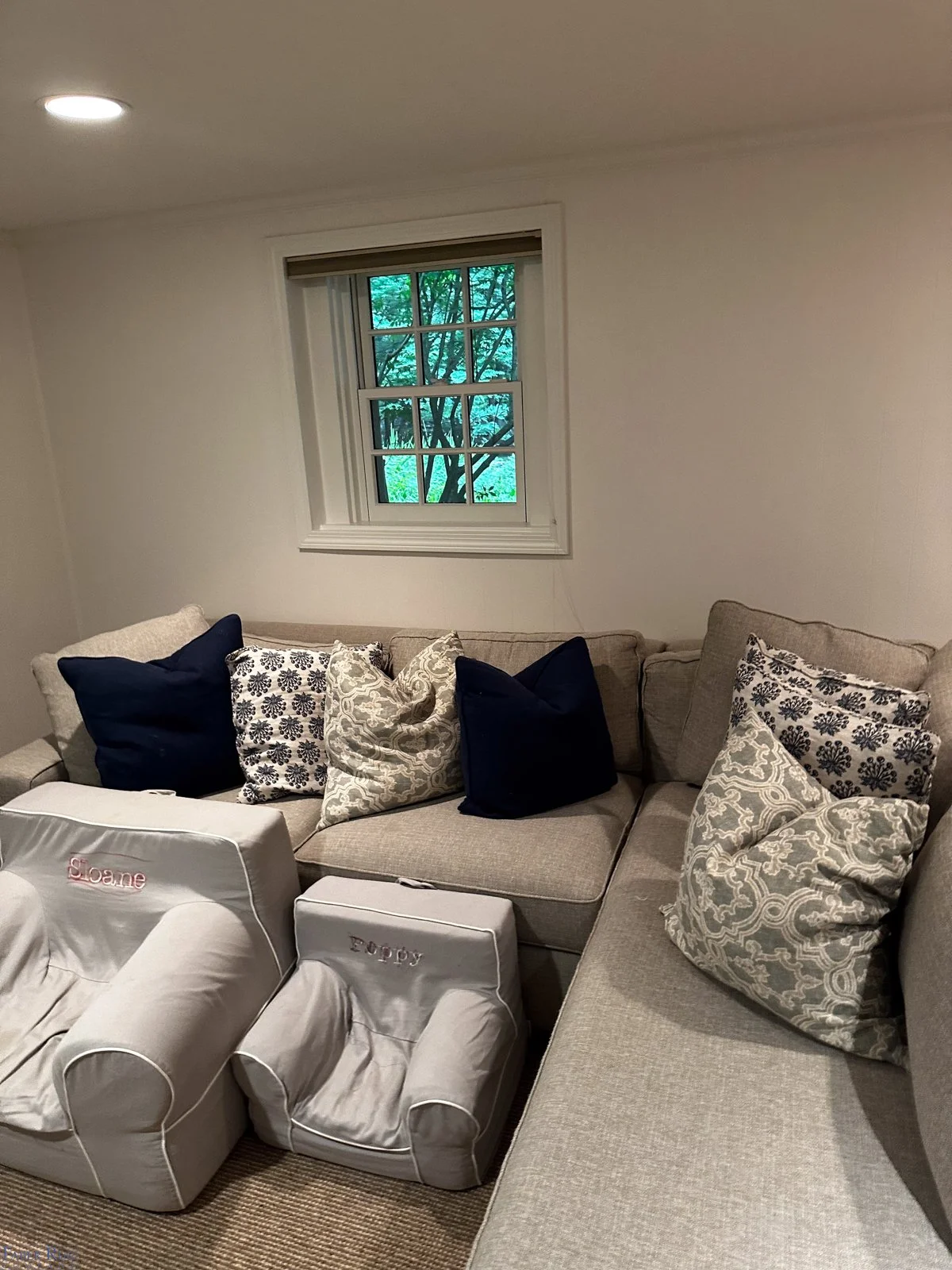 Living room with beige sectional sofa, navy and patterned pillows, a window with trees outside, and two small white footstools labeled 'Sloane' and 'Poppy' in front of the sofa.