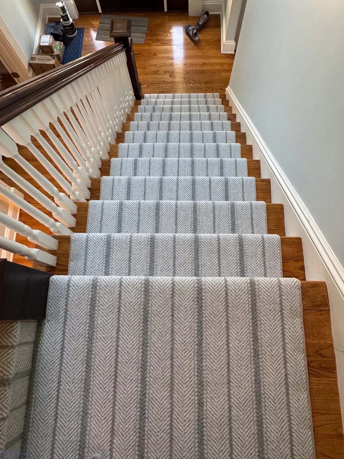 Top-down view of a carpeted staircase with a striped gray runner, wooden handrail, and white spindles, leading to a hardwood floor landing with shoes and boxes.