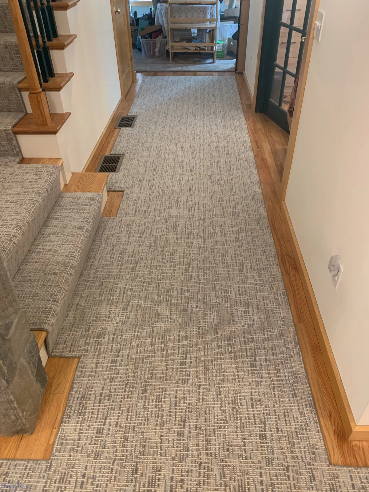 A hallway with a patterned beige and gray rug, wooden stairs on the left, and a glass-paneled door on the right. The hallway opens to a room with shelves and storage containers at the far end.