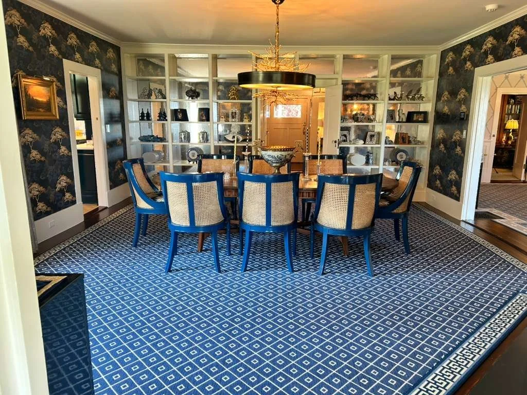 A dining room with a wooden table surrounded by ten blue and beige chairs, patterned blue rug, dark floral wallpaper, built-in white shelving with decorations, and a gold chandelier hanging above the table.