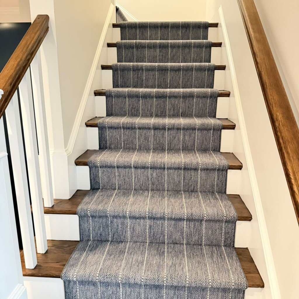 Carpeted staircase with a gray and white striped pattern, wooden steps, white risers, and wooden handrails on both sides.