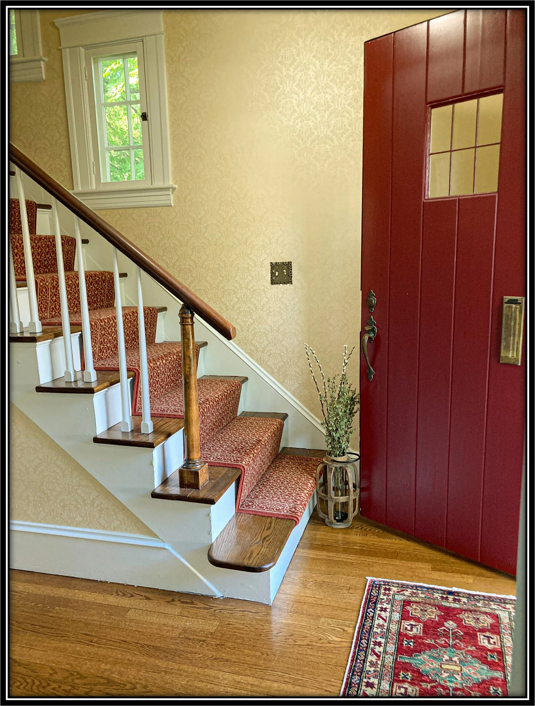 Interior of a home's entryway with a staircase carpeted in red and gold, a red front door with glass panels, a small window with green foliage outside, a vase with decorative sticks or branches, a patterned rug on hardwood floor, and beige wallpaper 