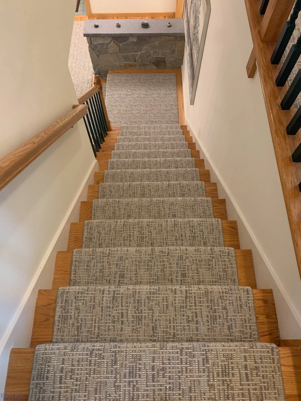 Top-down view of a staircase with a beige carpet runner, wooden steps, and a black metal handrail on one side, leading to a small landing area with a stone wall and a carpeted floor.