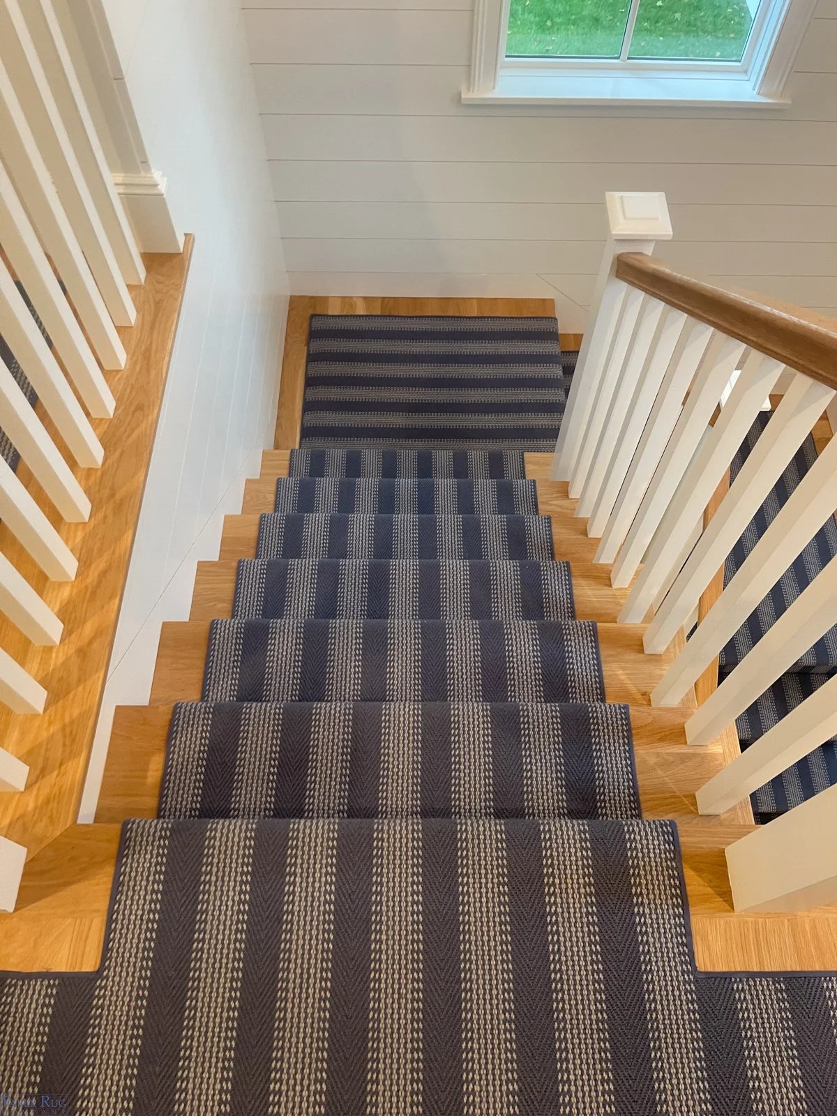 View of a staircase with carpeted steps and a blue, tan, and white striped runner. The staircase has a wooden handrail and white spindles, with a window at the top showing green grass outside.
